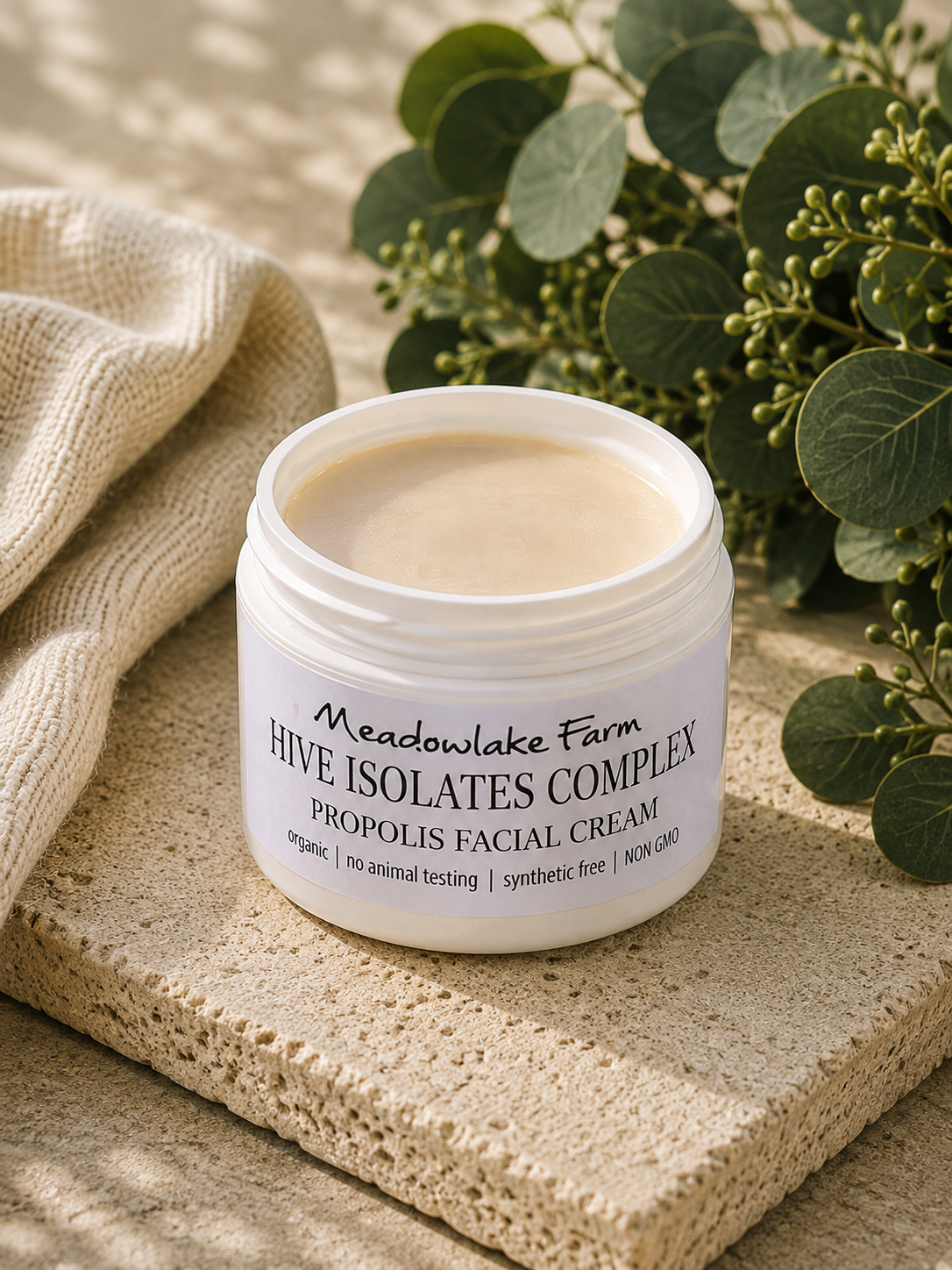 A jar of Meadowlake Farm Hive Isolates Complex Propolis Facial Cream sits on a textured stone slab next to a beige knit fabric and green eucalyptus leaves.