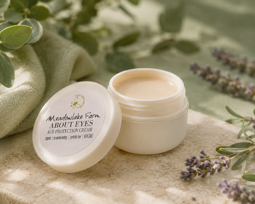 An open white cosmetic jar of Meadowlake Farm About Eyes Age Protection Cream sits on a stone surface with its lid beside it. The scene is styled with green leaves, a folded cloth, and lavender sprigs in soft natural light.