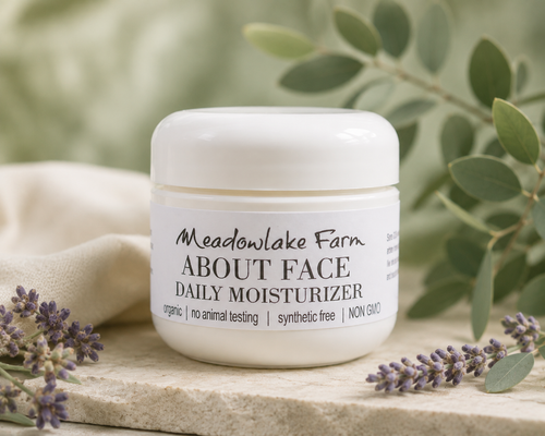 A white jar of Meadowlake Farm About Face Daily Moisturizer sits on a light stone surface with soft green leaves and small purple flowers around it.