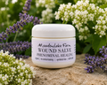 A jar of Meadowlake Farm Wound Salve set on a stone surface with purple flowers and white blossoms in the background.