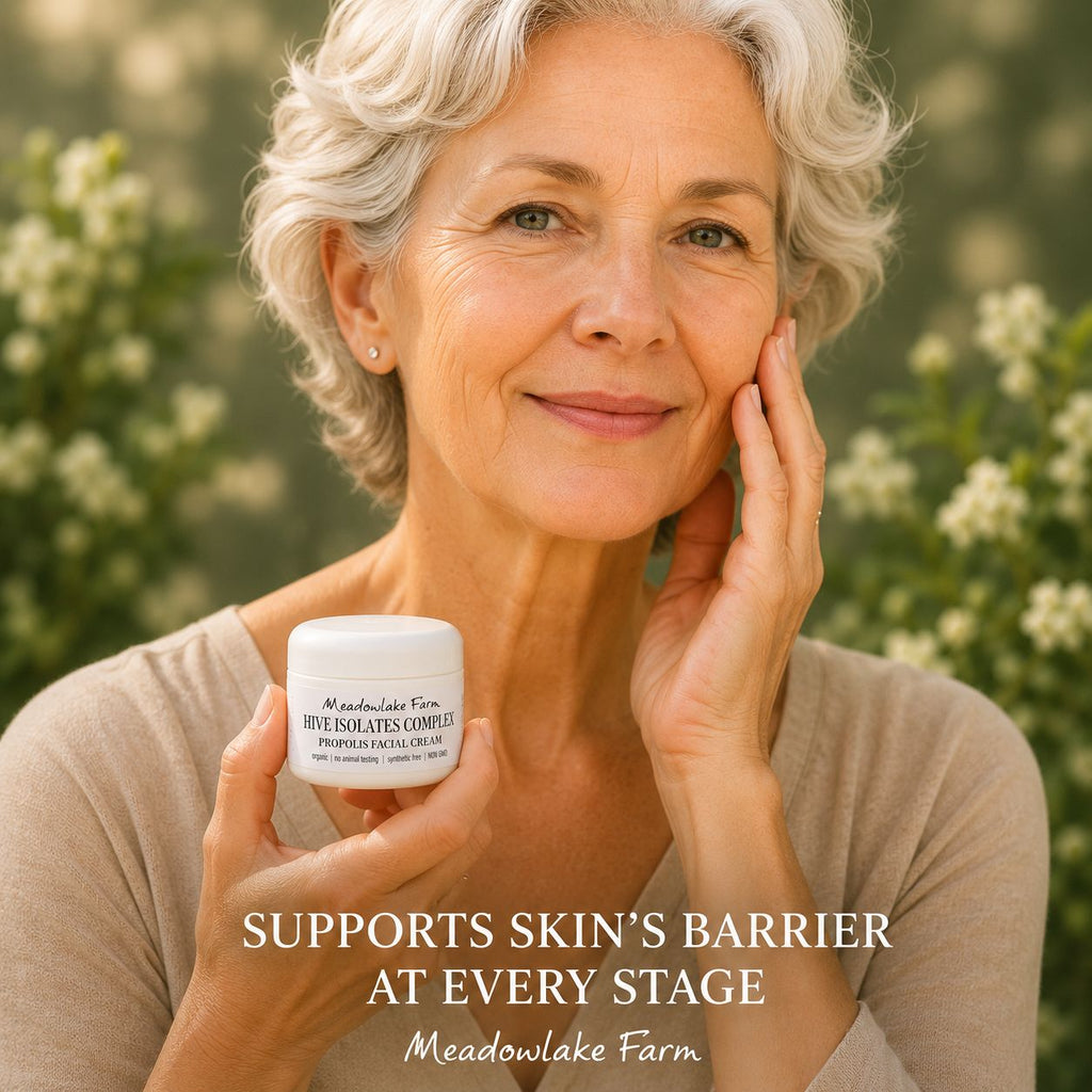 A smiling older woman with short gray hair holds a white jar of Meadowlake Farm Hive Isolates Complex Hydration Cream against a soft-focus outdoor background of greenery and white flowers. Text overlays read 'SUPPORTS SKIN'S BARRIER AT EVERY STAGE' and 'Meadowlake Farm'.